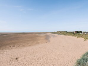 Une plage de sable fin, un ciel dégagé, quelques bâtiments visibles au loin et une zone herbeuse délimitée par une clôture.