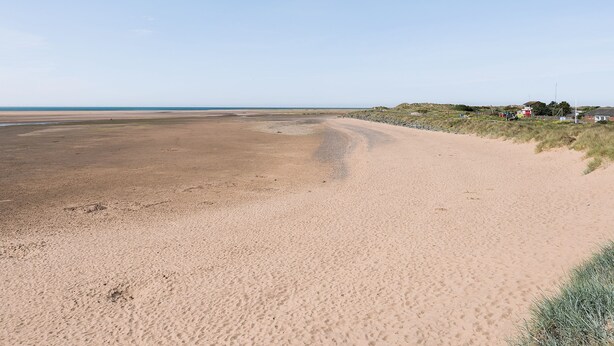 Une plage de sable fin, un ciel dégagé, quelques bâtiments visibles au loin et une zone herbeuse délimitée par une clôture.