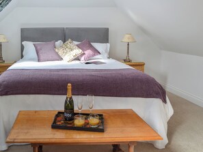 Una cama bien hecha con cabecera gris, ropa de cama blanca y morada, y una botella de champán sobre una bandeja con dos copas.