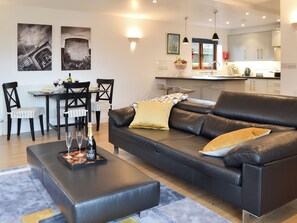 A modern living room with a black leather sofa, a dining table with chairs, and a kitchen in the background. - 1 bedroom accommodation in Windmill Hill, near Hailsham (Windmill Hill, near Hailsham)