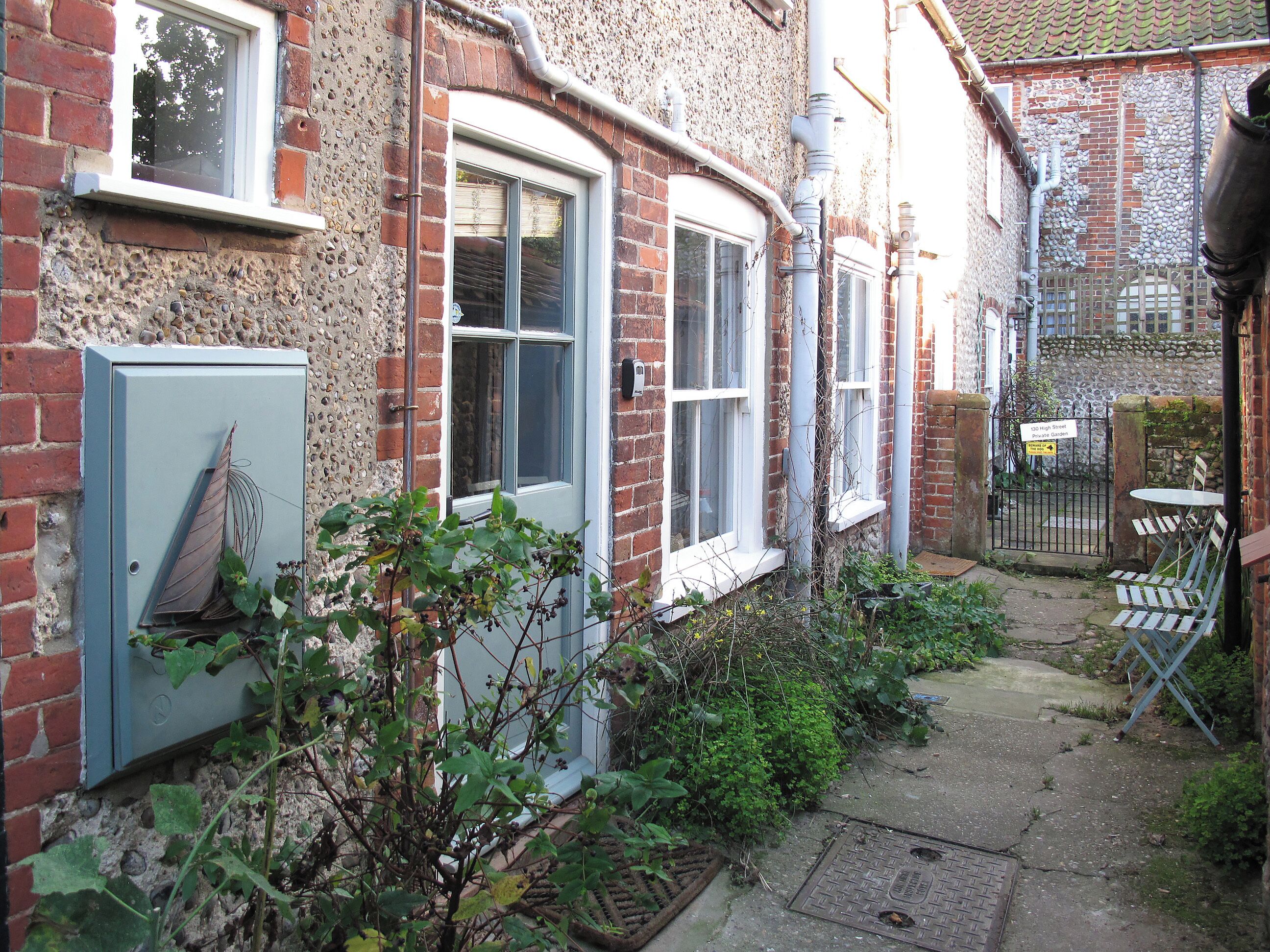 A narrow alleyway with a brick building, a metal utility box, and a small garden area with a table and chairs.