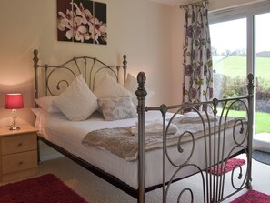 Une chambre à coucher avec un lit en métal, du linge de lit blanc, une lampe rouge, une petite table de chevet et une vue sur un jardin, visible à travers une fenêtre ornée de rideaux à motifs floraux.