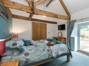 Een slaapkamer met een houten plafond, een bed met bloemenbeddengoed, een nachtkastje, een televisie en een schuifdeur naar buiten.