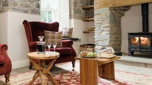 A cozy living room with a stone fireplace, a red armchair, a wooden coffee table, and a stone wall. - Waterfall Wood Cottage (Glenridding, by Ullswater)