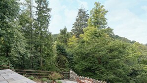A stone patio with a wooden table and chairs, surrounded by a stone wall and a forested hillside. - Waterfall Wood Cottage (Glenridding, by Ullswater)