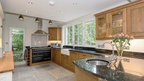 A modern kitchen with wooden cabinets, a built-in oven, a microwave, and a large window providing natural light. - Waterfall Wood Cottage (Glenridding, by Ullswater)