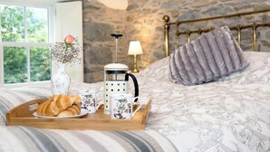 A bed with a stone wall, a bedside lamp, and a tray with croissants and a coffee maker. - Waterfall Wood Cottage (Glenridding, by Ullswater)