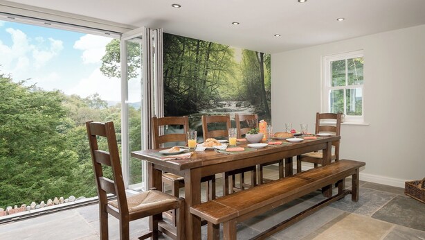 A dining area with a table set for four, surrounded by wooden chairs and benches, with a scenic forest view through large windows. - Waterfall Wood Cottage (Glenridding, by Ullswater)