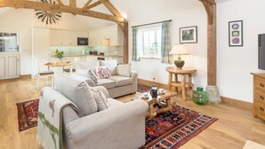 A spacious living area with a high ceiling, wooden beams, a sofa, a coffee table, and a kitchen in the background. - The Old Dairy-UKC2112 (Royal Wootton Bassett)