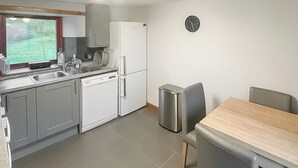 Una cuina compacta amb armaris grisos, un rentaplats blanc, un frigorífic blanc, un cendrer de metall inoxidable i una taula de dinar de fusta amb dues cadires.