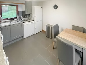 A compact kitchen with grey cabinets, a white dishwasher, a white refrigerator, a stainless steel trash bin, and a wooden dining table with two chairs.