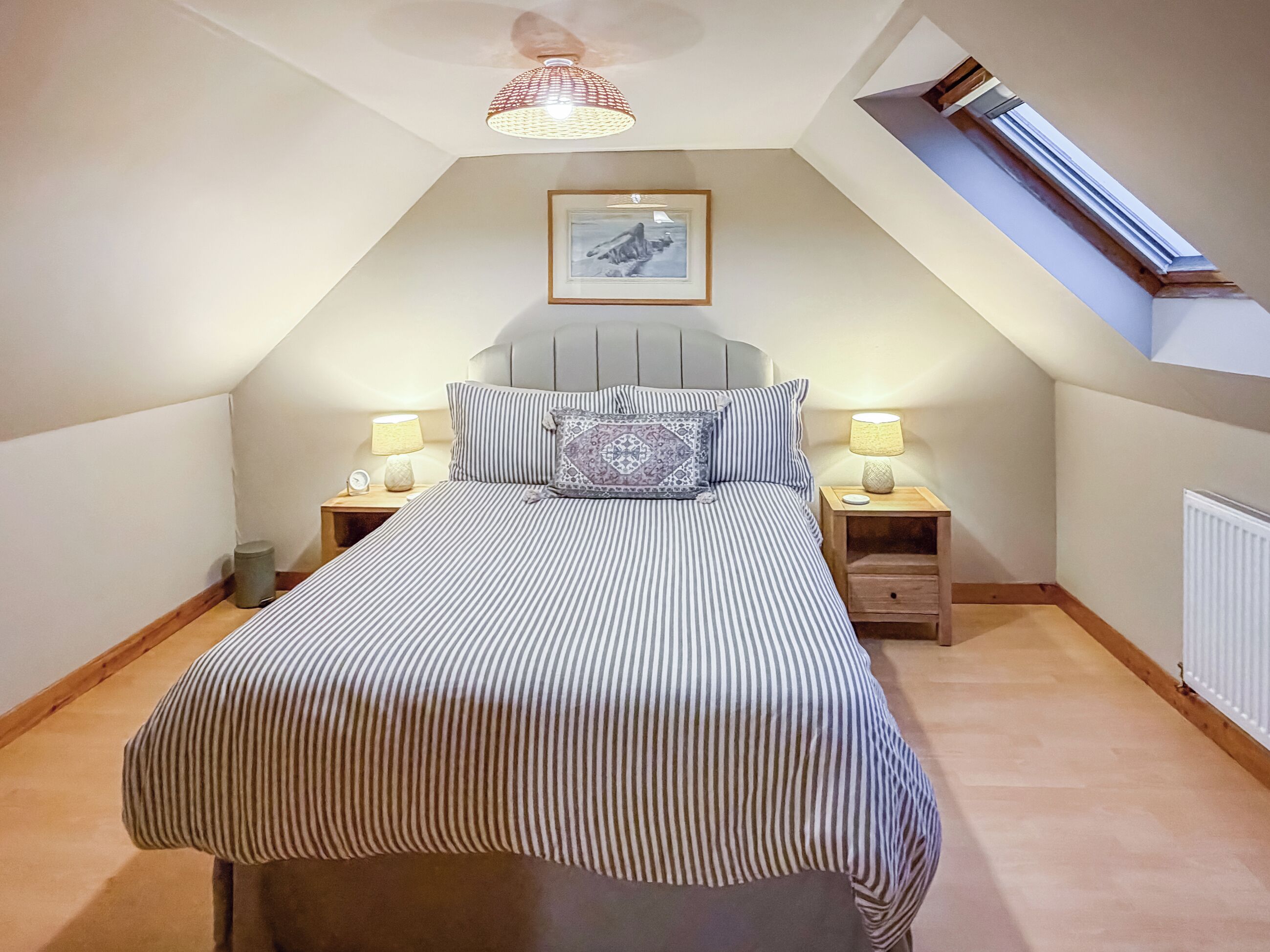 A bedroom with a sloped ceiling, a bed with striped bedding, two bedside lamps, a framed picture on the wall, and a window with a view.