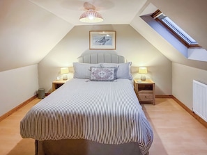 A bedroom with a sloped ceiling, a bed with striped bedding, two bedside lamps, a framed picture on the wall, and a window with a view.