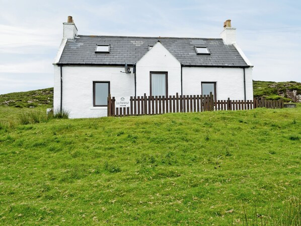 A white house with a dark roof, a wooden fence, and a grassy hill in the background.