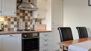 A kitchen with white cabinets, a gas stove, an oven, and a tiled backsplash. There is a wooden cutting board and a knife on the counter. - 2 bedroom accommodation in Allonby, near Maryport (Allonby, near Maryport)