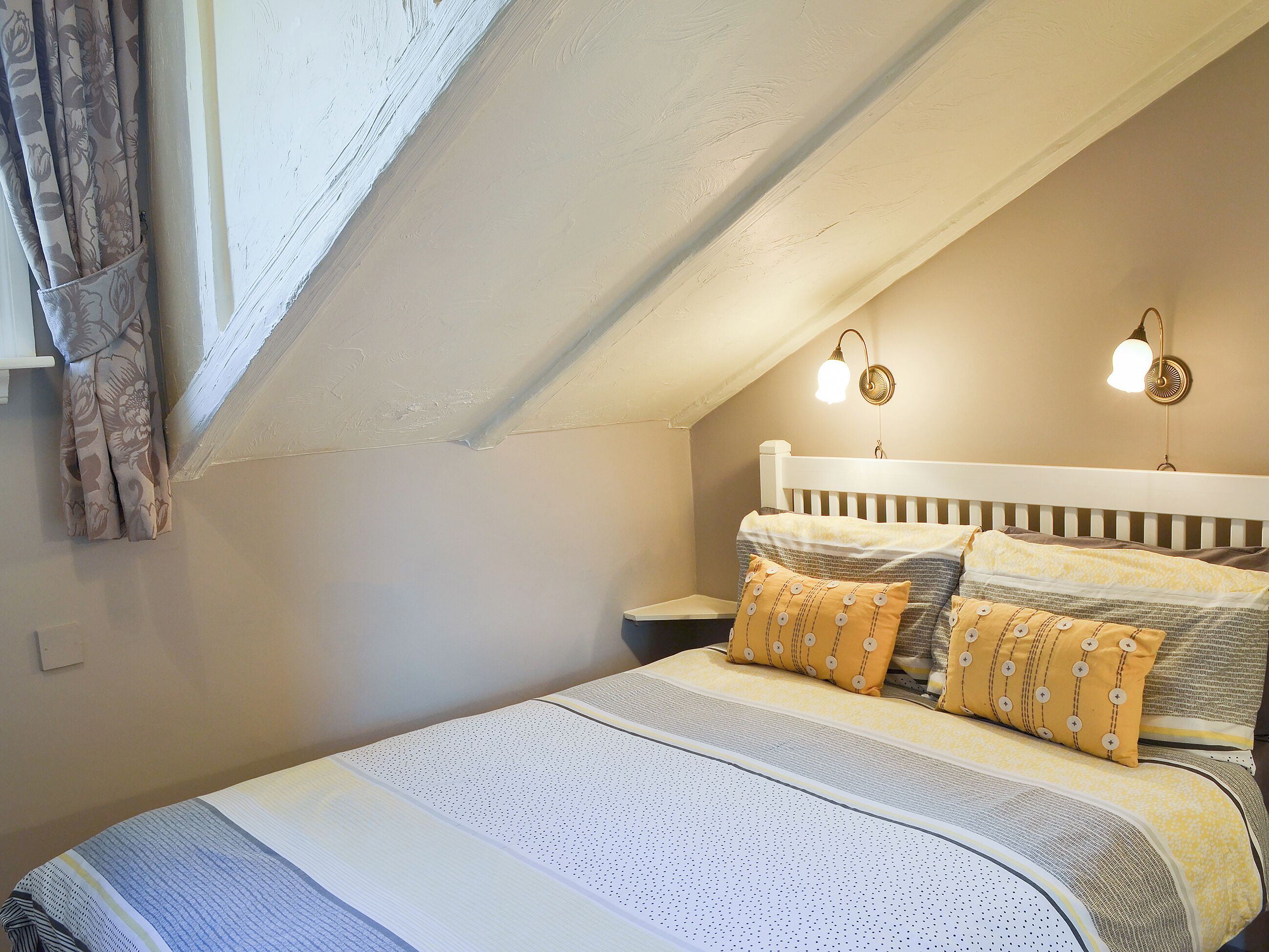Een slaapkamer met een schuine kap, een bed met beddengoed met een patroon, twee wandlampen en een raam met gordijnen.
