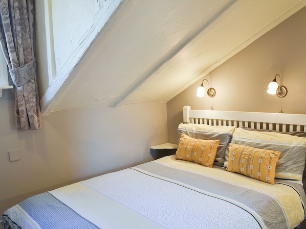A bedroom with a sloped ceiling, a bed with patterned bedding, two wall-mounted lamps, and a window with curtains.
