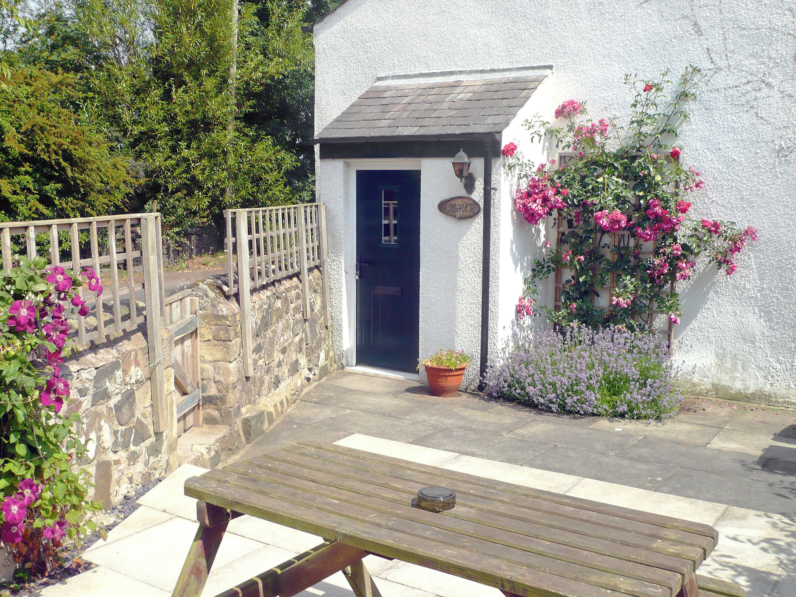 A small white cottage with a dark blue door, a wooden fence, and a garden with pink flowers.