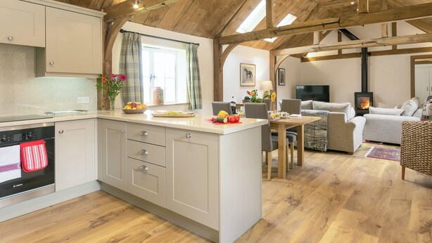 A spacious kitchen with wooden beams, a dining area with a table and chairs, and a living room with a sofa and fireplace. - The Calf Shed-UKC2113 (Royal Wootton Bassett)