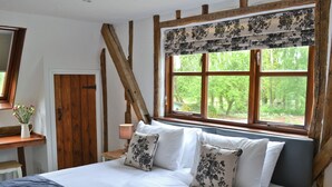 A bedroom with a bed, wooden beams, a window with a floral valance, a bedside table with a lamp, and a vase with flowers.