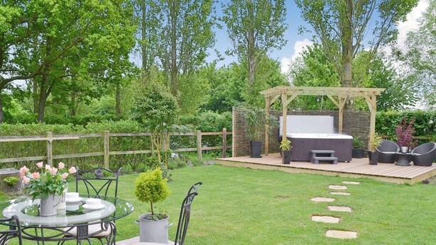 A patio with a round glass table, metal chairs, a small potted plant, and a wooden pergola with a hot tub.