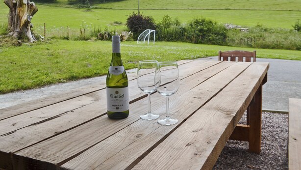 A wooden table with a bottle of wine and two glasses, set against a backdrop of a green field and trees. - 4 bedroom accommodation in Capel Bangor, near Aberystwyth (Capel Bangor, near Aberystwyth)