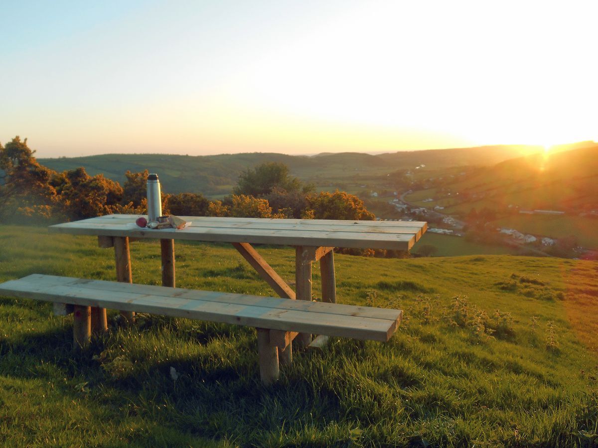 Une table de pique-nique est installée sur une colline verdoyante, offrant une vue panoramique sur des collines ondulantes et un coucher de soleil en arrière-plan.
