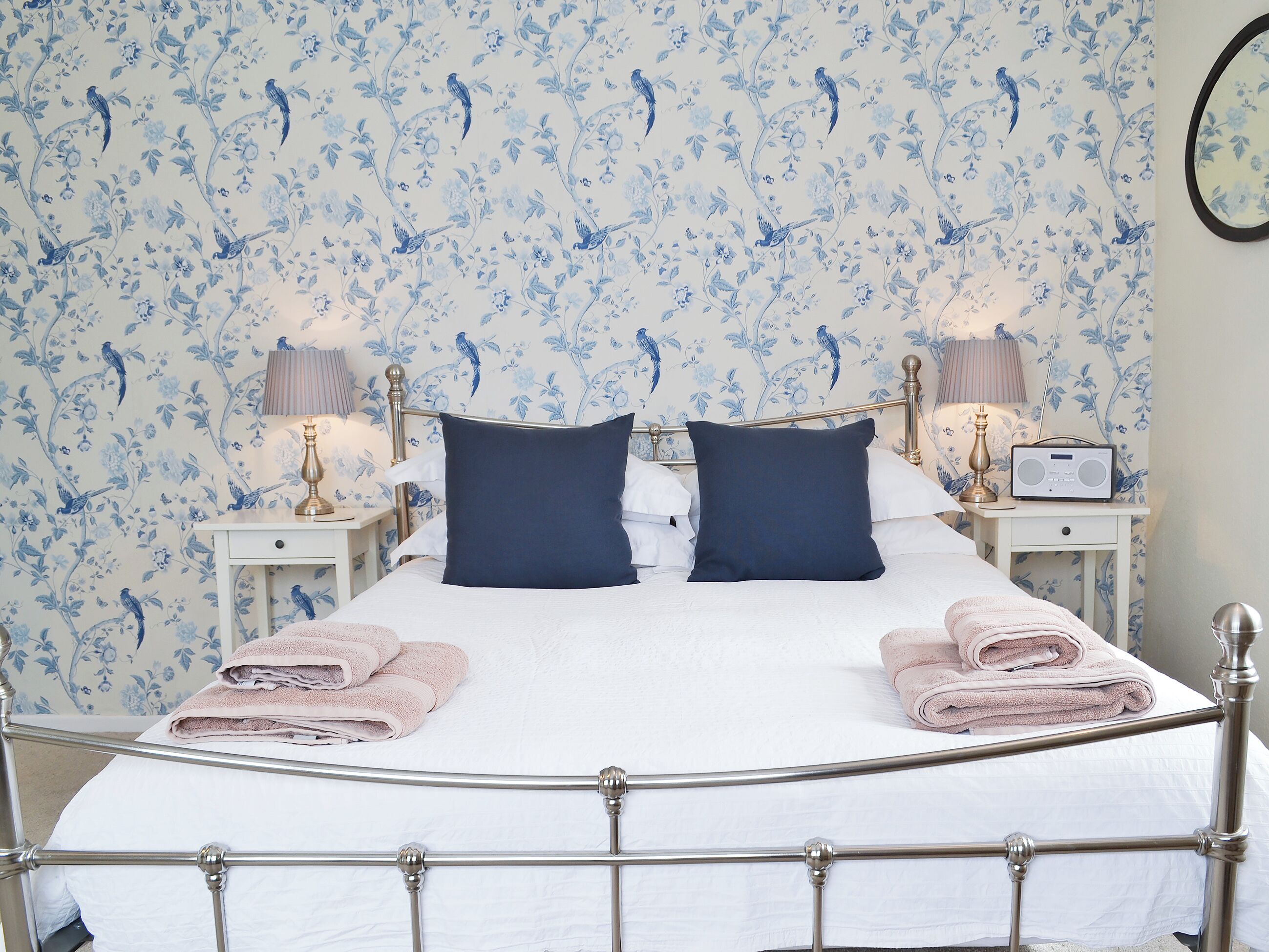 A neatly made bed with two pillows, a bedside table with a lamp and radio, and a wall with floral wallpaper.