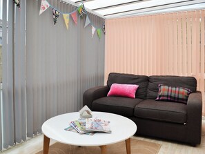 A living room with a brown sofa, a white coffee table with magazines, and a window with blinds. - 2 bedroom accommodation in Mundesley, near North Walsham (Mundesley, near North Walsham)