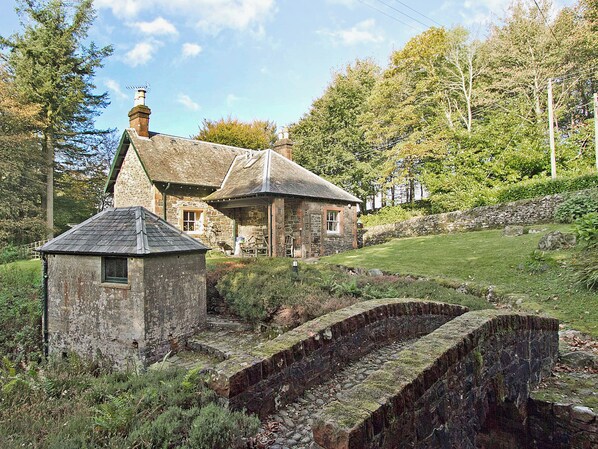 A stone house with a traditional design, a small adjacent structure, and a stone bridge in the foreground.