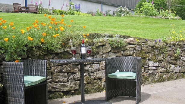 An outdoor seating area with wicker chairs and a table, a stone wall, and a grassy lawn with flowers. - Attractive farm cottage with period features and views over fields and fells. (Caldbeck, near Keswick)