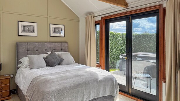 A bedroom with a large bed, a wooden ceiling beam, two framed pictures on the wall, and a sliding glass door leading to an outdoor patio. - 1 bedroom accommodation in White Colne, near Colchester (White Colne, near Colchester)