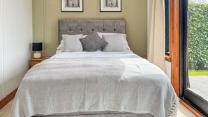 A bedroom with a large bed, a nightstand, a lamp, and a view of greenery outside. - 1 bedroom accommodation in White Colne, near Colchester (White Colne, near Colchester)