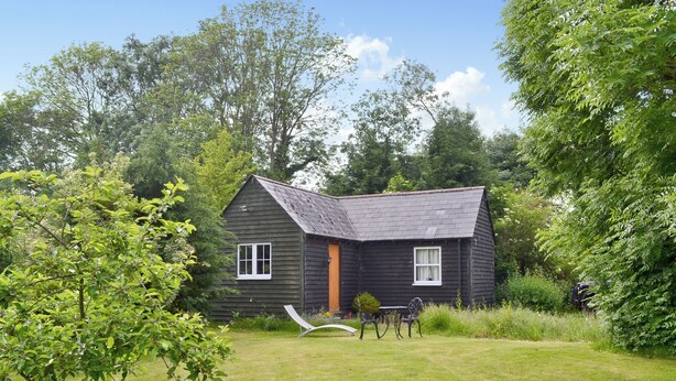 A small, dark wooden house with a sloped roof, surrounded by a grassy lawn and dense trees. - 1 bedroom accommodation in Burgate, near Fordingbridge (Burgate, near Fordingbridge)