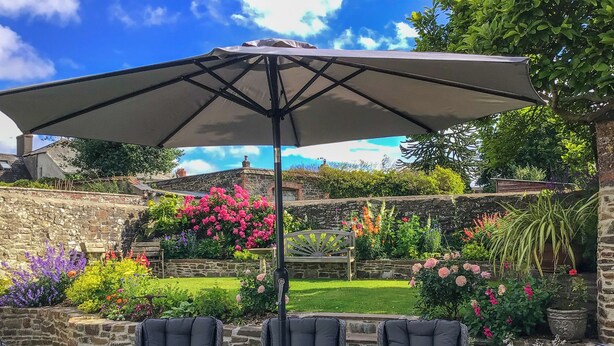 A patio with a glass table and chairs, an umbrella, and a stone wall with flowering plants. - 3 bedroom accommodation in Chulmleigh, near South Molton (Chulmleigh, near South Molton)