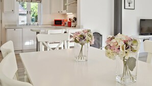 A modern kitchen with white cabinets, a red microwave, and two vases with flowers on the dining table. - Wishing Well Cottage (Mathon, near Malvern)
