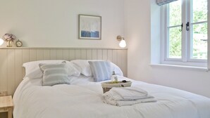 A neatly made bed with pillows and a tray of towels, a bedside table with a lamp, and a window with curtains. - Wishing Well Cottage (Mathon, near Malvern)