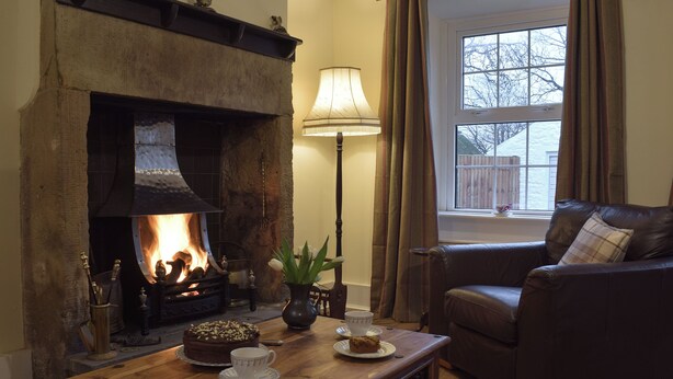 A cozy living room with a fireplace, a leather sofa, a wooden coffee table with cakes, and a window with curtains.