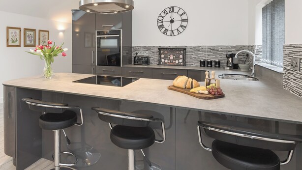 A modern kitchen with a central island, bar stools, a clock, and a tiled backsplash. - 1 bedroom accommodation in Boughton, near Downham Market (Boughton, near Downham Market)