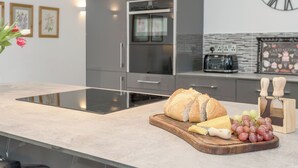 A modern kitchen with a built-in oven, a clock, and a cutting board with bread, cheese, and grapes. - 1 bedroom accommodation in Boughton, near Downham Market (Boughton, near Downham Market)