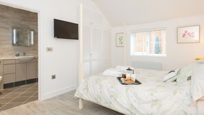 A bedroom with a bed, a bedside table with a tray of breakfast items, a window with blinds, a flat-screen TV, and a bathroom with a sink and mirror. - 1 bedroom accommodation in Boughton, near Downham Market (Boughton, near Downham Market)