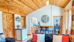 Een gezellige woonkamer met een zwarte leren bank, een houten plafond en een zichtbare keuken door een open deur.