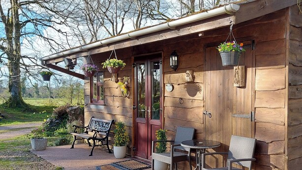 Een rustieke houten blokhut met een veranda, een bank, twee rieten stoelen en bloemenmanden.