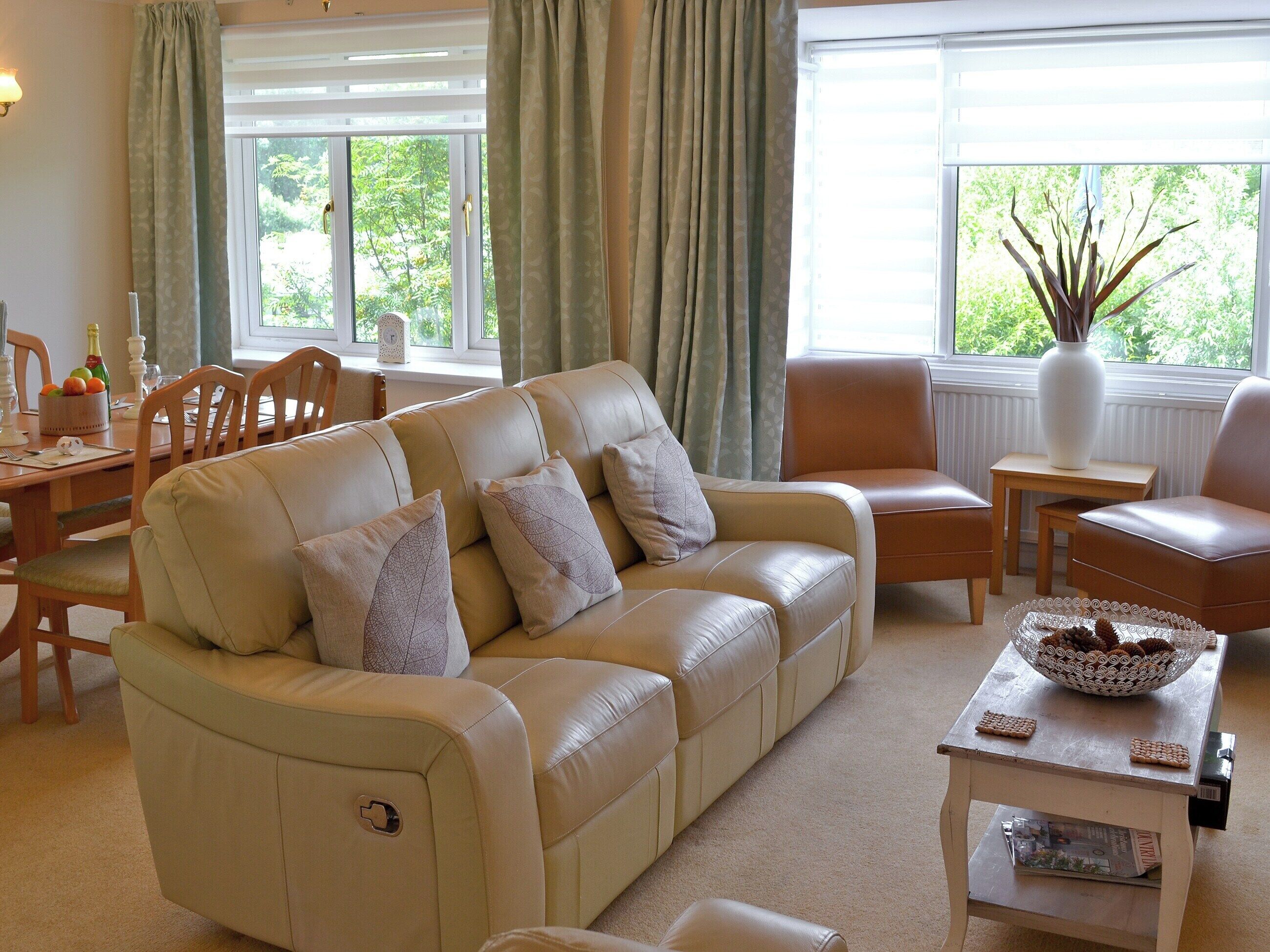 A living room with a beige sofa, two armchairs, a coffee table, and a dining table with chairs. There are windows with curtains, a vase on the windowsill, and a bowl of cookies on the coffee table.