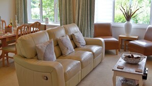 Ein Wohnzimmer mit einem beigefarbenen Sofa, zwei Sesseln, einem Couchtisch und einem Esstisch mit Stühlen. Es gibt Fenster mit Vorhängen, eine Vase auf der Fensterbank und eine Schale Kekse auf dem Couchtisch.