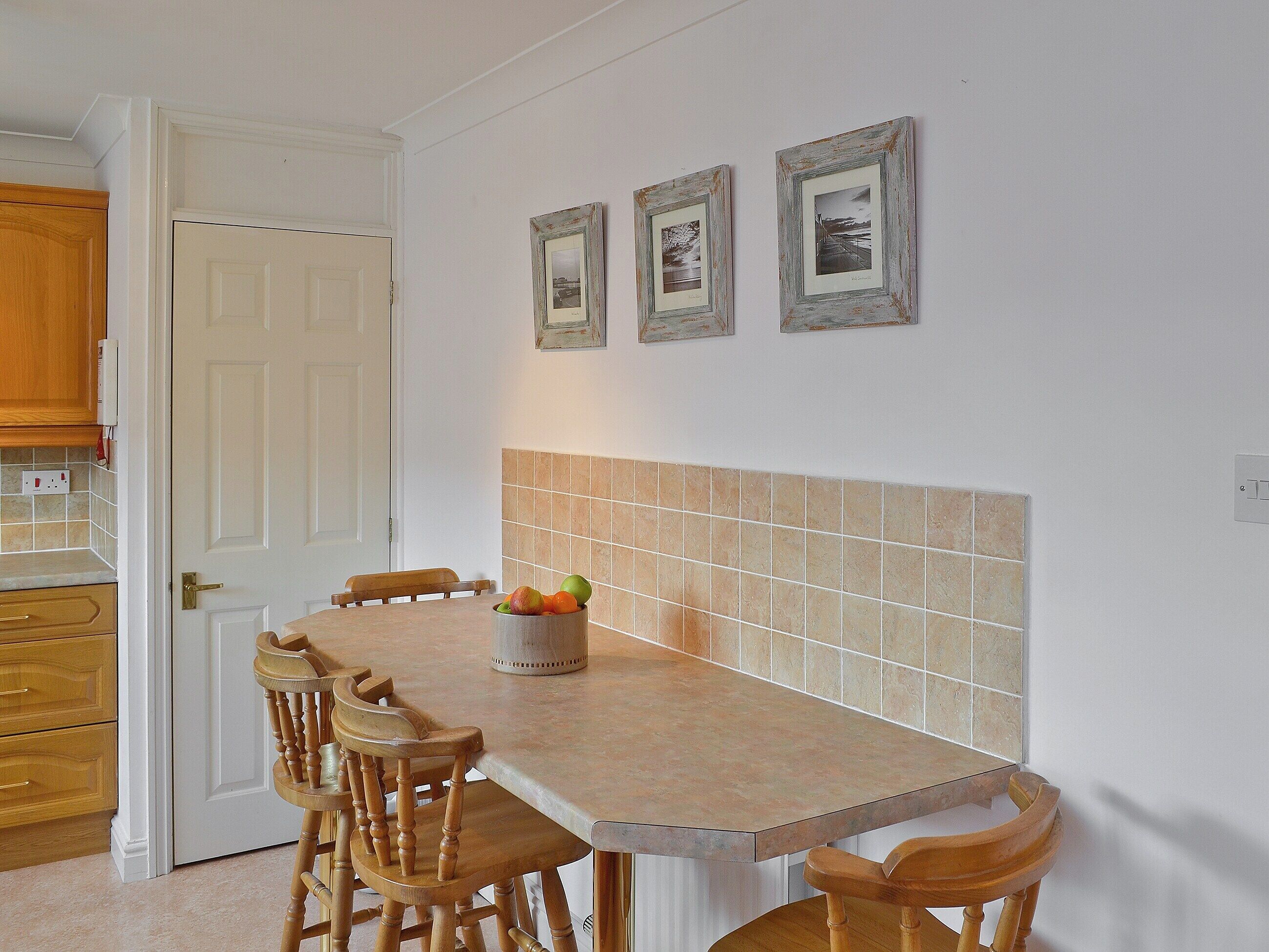 A kitchen with a tiled backsplash, wooden chairs, a fruit bowl on the table, and framed pictures on the wall.
