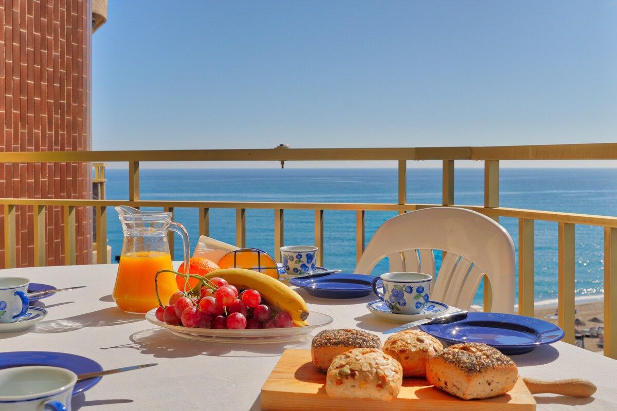 A table set with breakfast items including bread, fruit, and juice, overlooking the sea.
