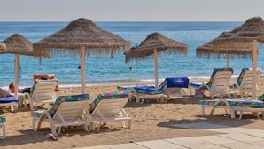 Ein Strand mit Reetdachschirmen und Liegestühlen, ein klares blaues Meer und einige Menschen im Wasser.