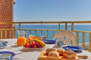 Una mesa dispuesta con alimentos para el desayuno, como uvas, un plátano y pasteles, con vista a un mar azul y despejado.