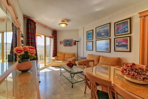 A living room with a glass coffee table, two sofas, a dining table with chairs, and a large window with curtains.